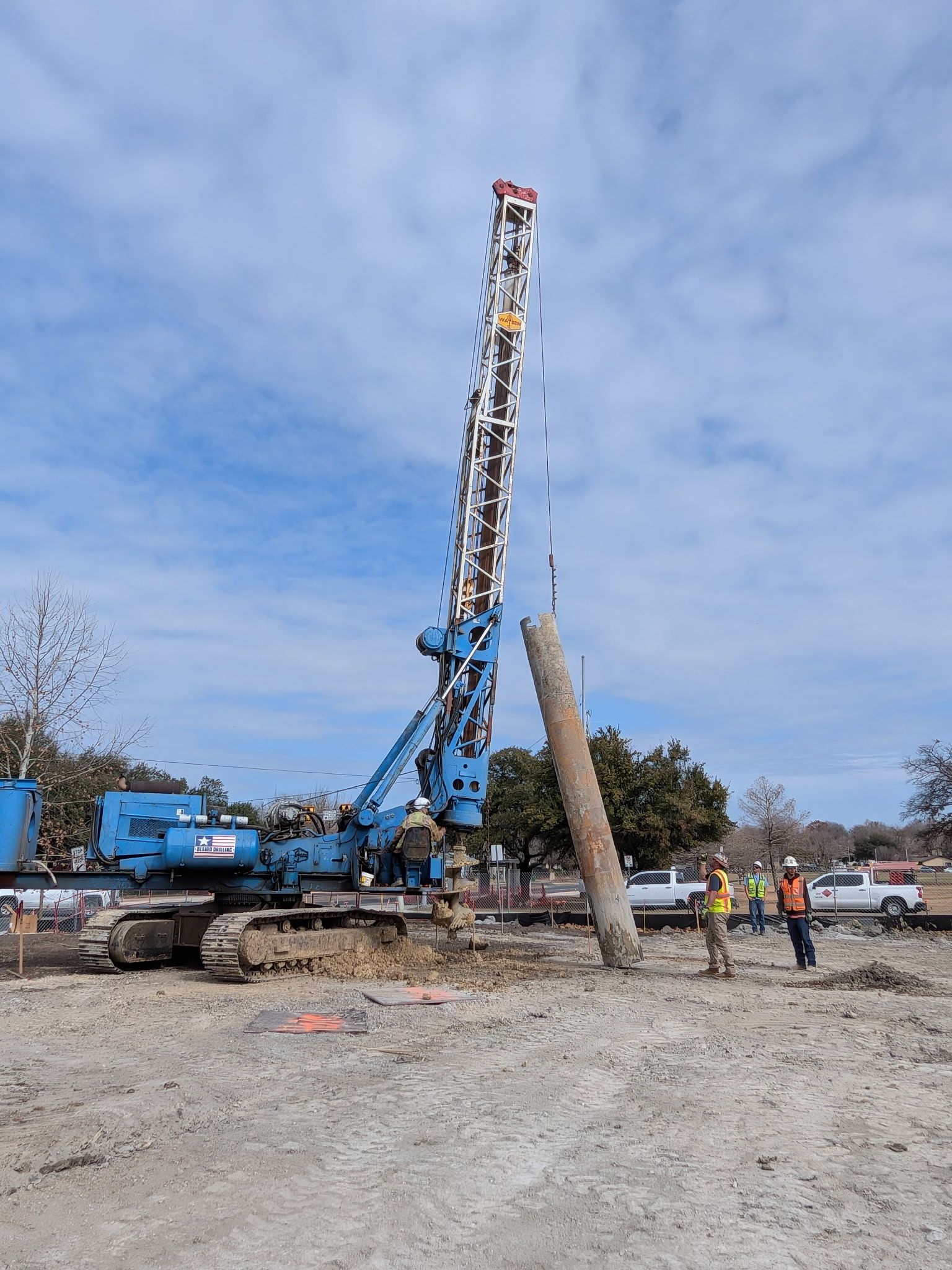 Blue crane lifting a tilted utility pole at a construction site under a cloudy sky
