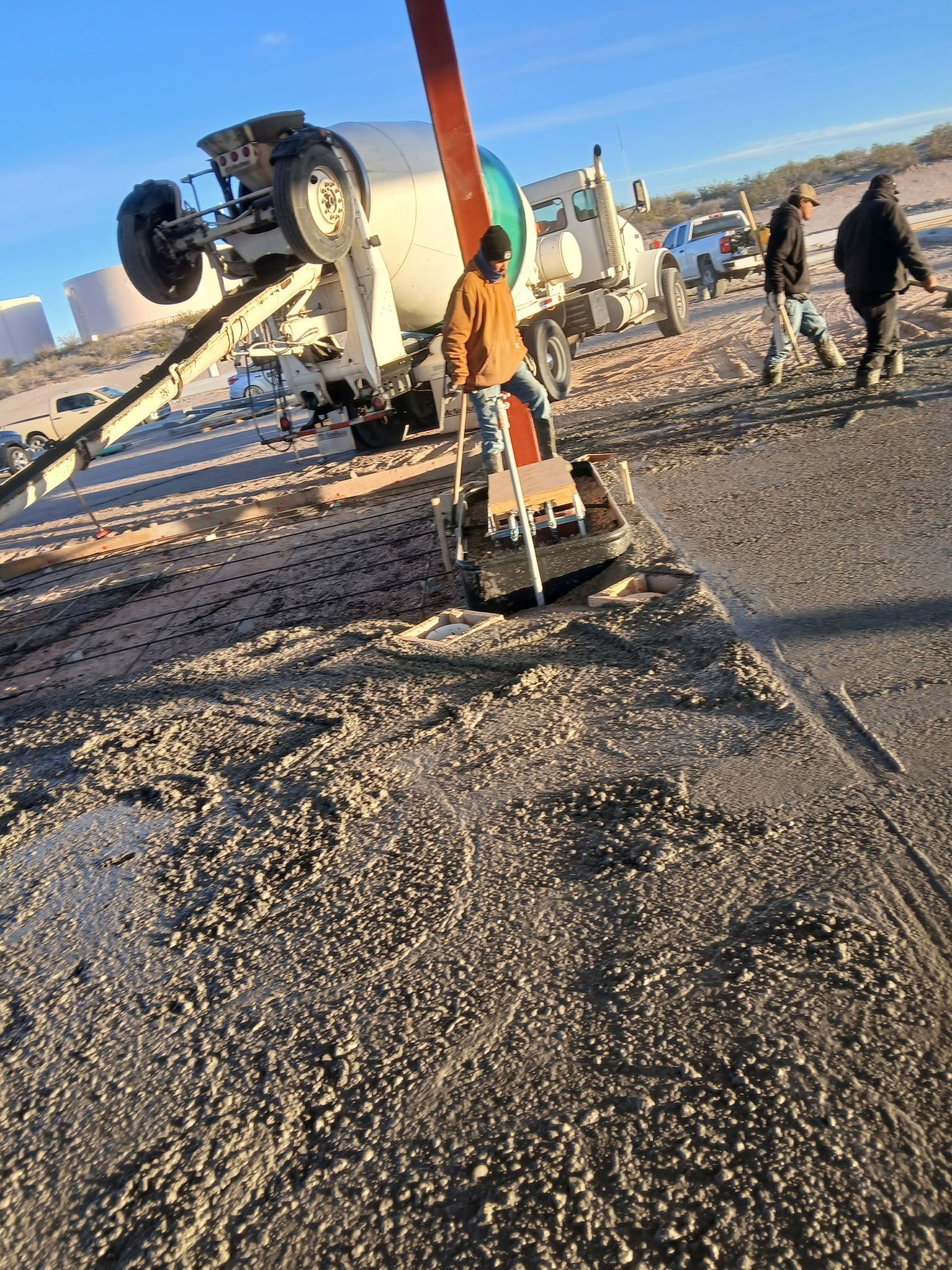 Construction equipment on a gravel site, with a crane lifting a large pipe at sunset.