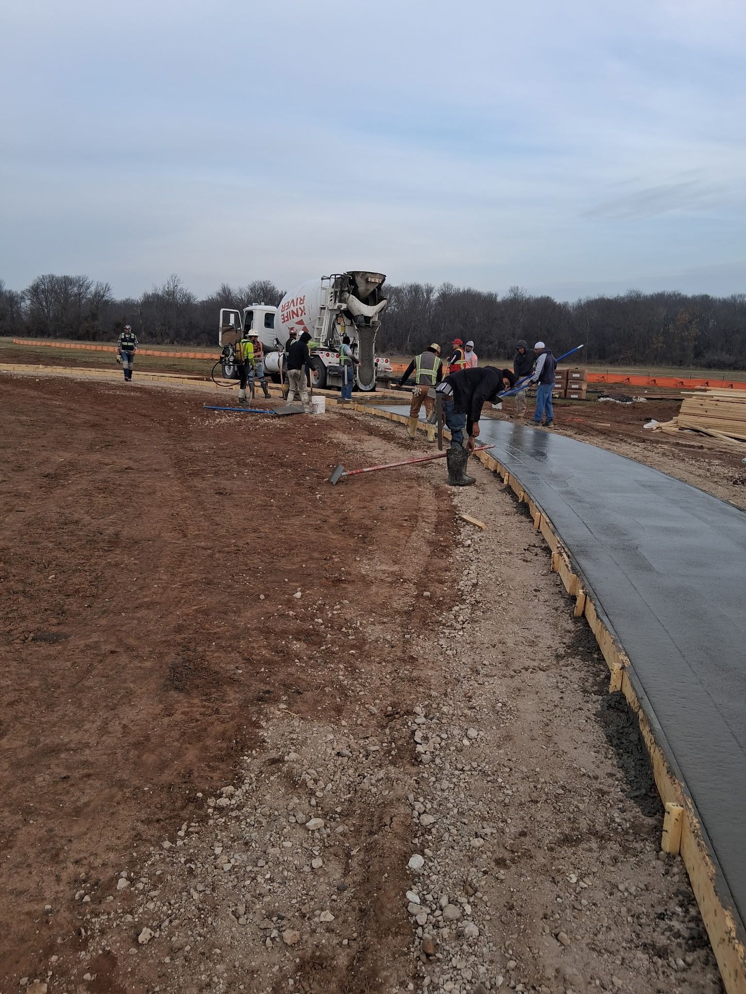 Workers near a concrete truck paving a road beside a muddy dirt lot under an overcast sky