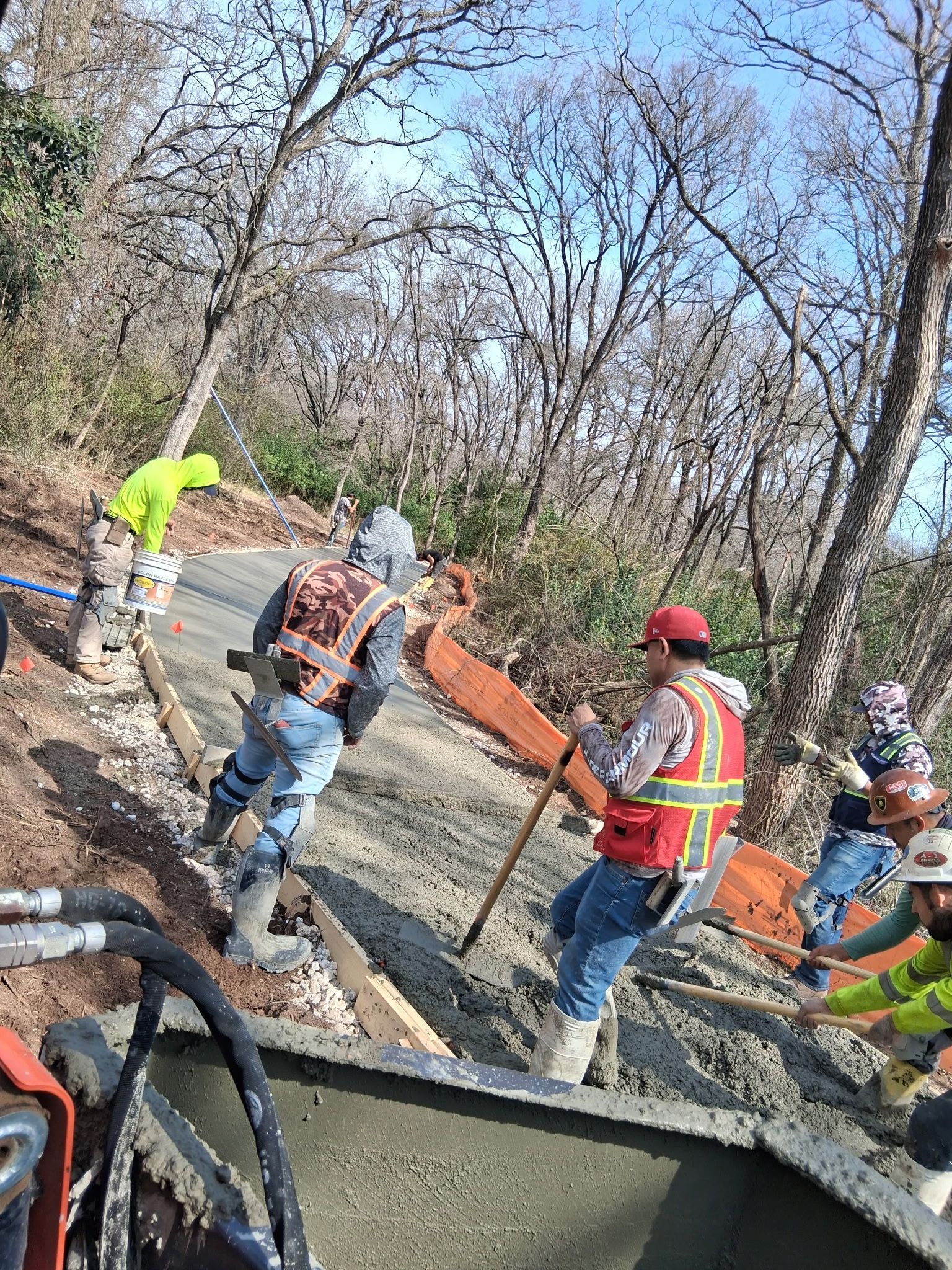 Road crew in safety vests repairing a gravel path beside wooded trees, with a backhoe nearby.
