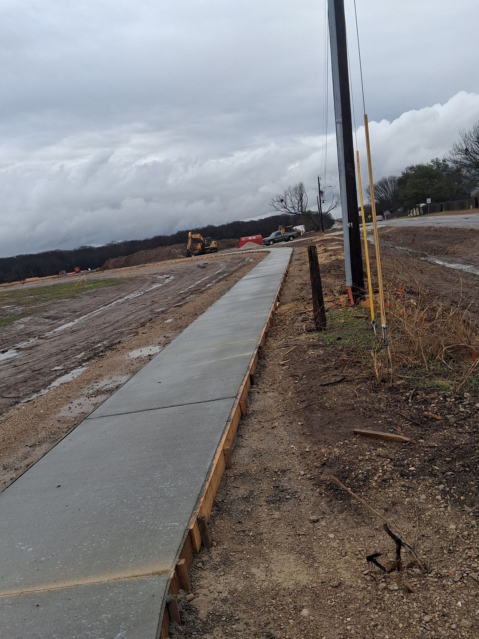 Concrete sidewalk through muddy construction site under cloudy sky, with utility poles along the right side.