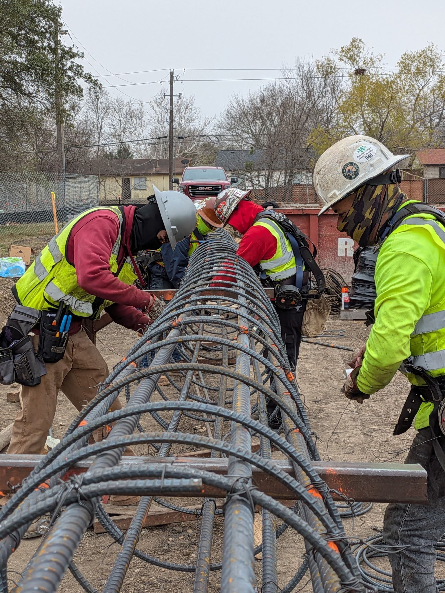 Construction workers in hard hats bend over rebar at a muddy outdoor job site