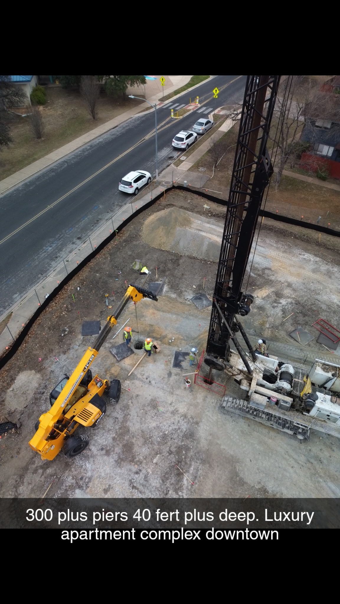 Construction site with yellow crane beside a road where several white cars drive past a downtown apartment complex