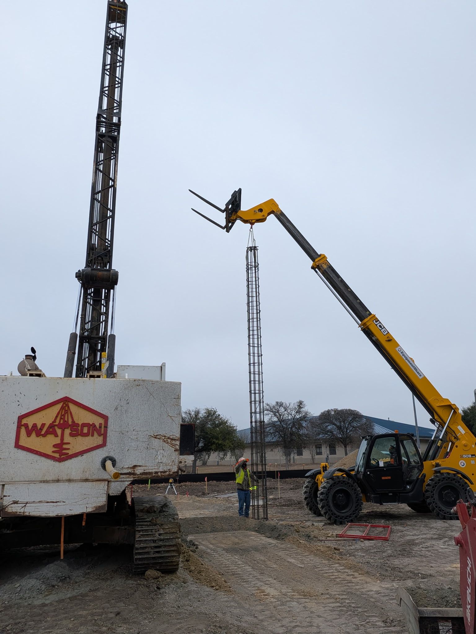 Construction site with a yellow crane lifting a tall lattice mast beside a truck trailer.