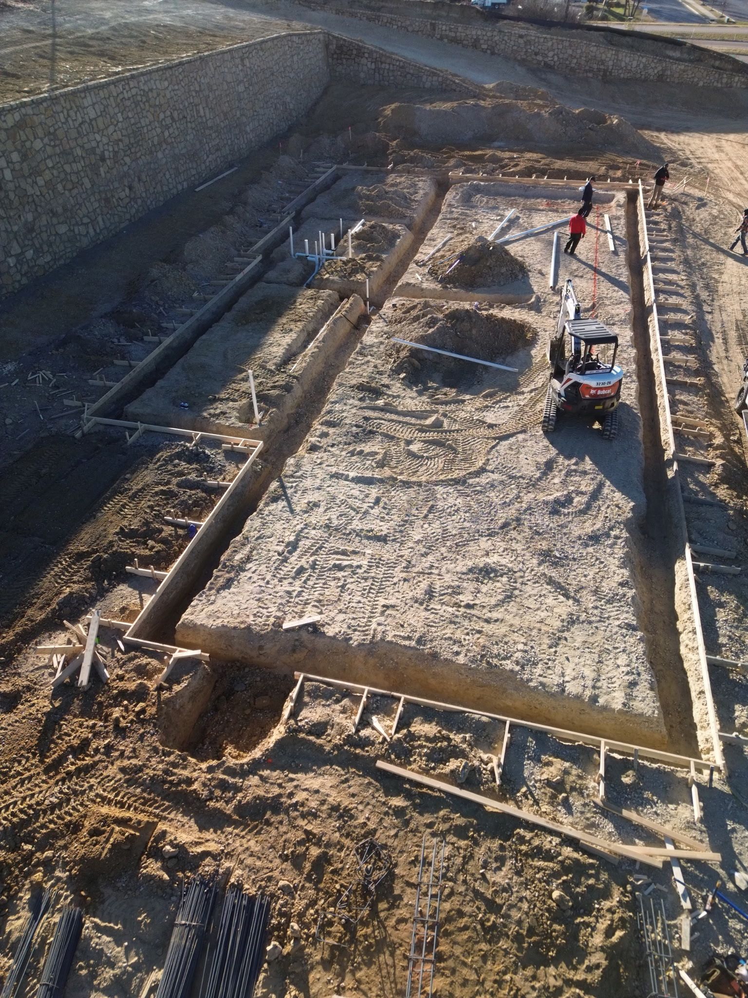 Aerial view of a rectangular excavation site with stone retaining walls, dirt floor, and two workers near equipment.