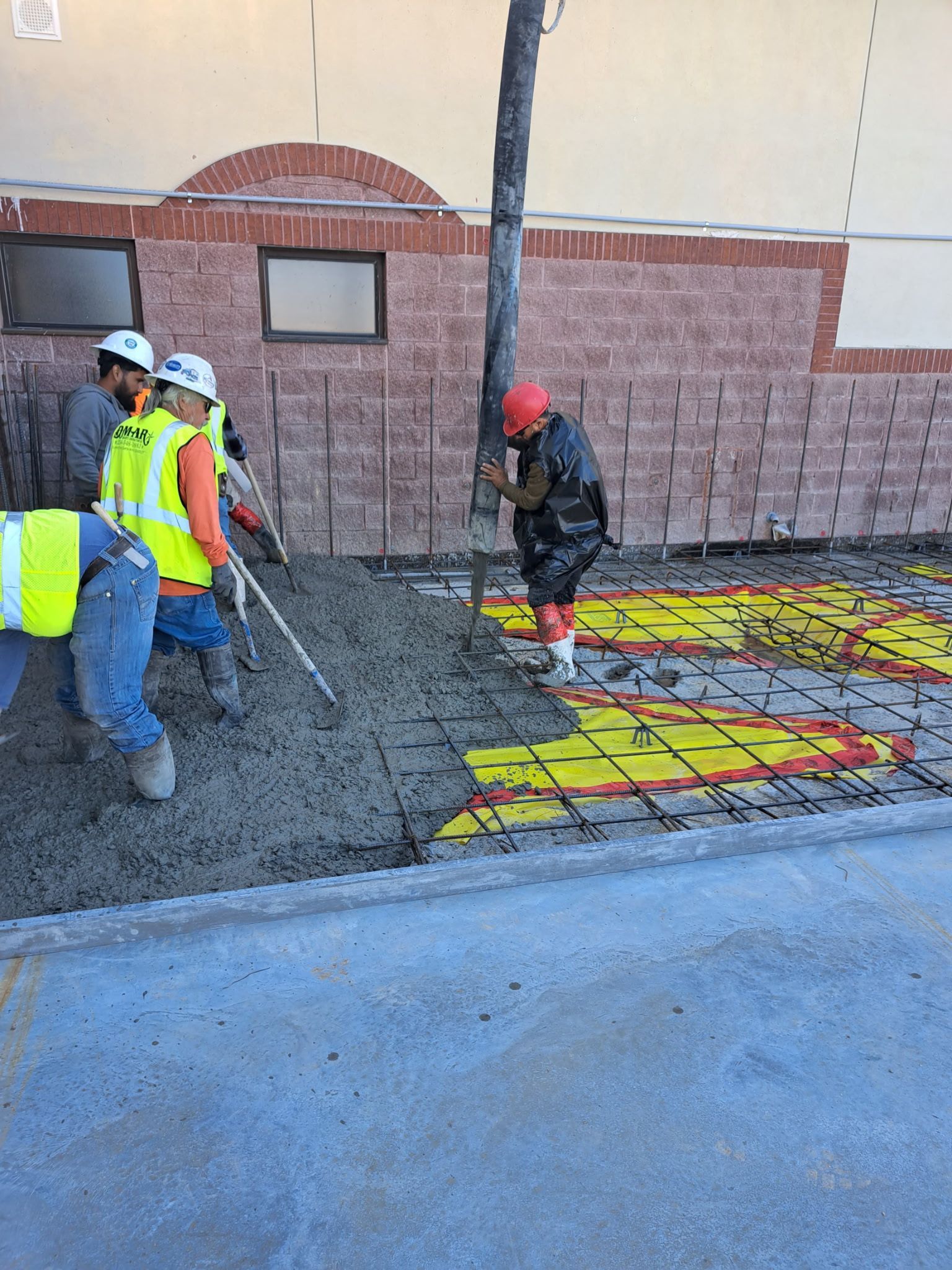 Workers laying concrete over a colorful patterned surface beside a brick building