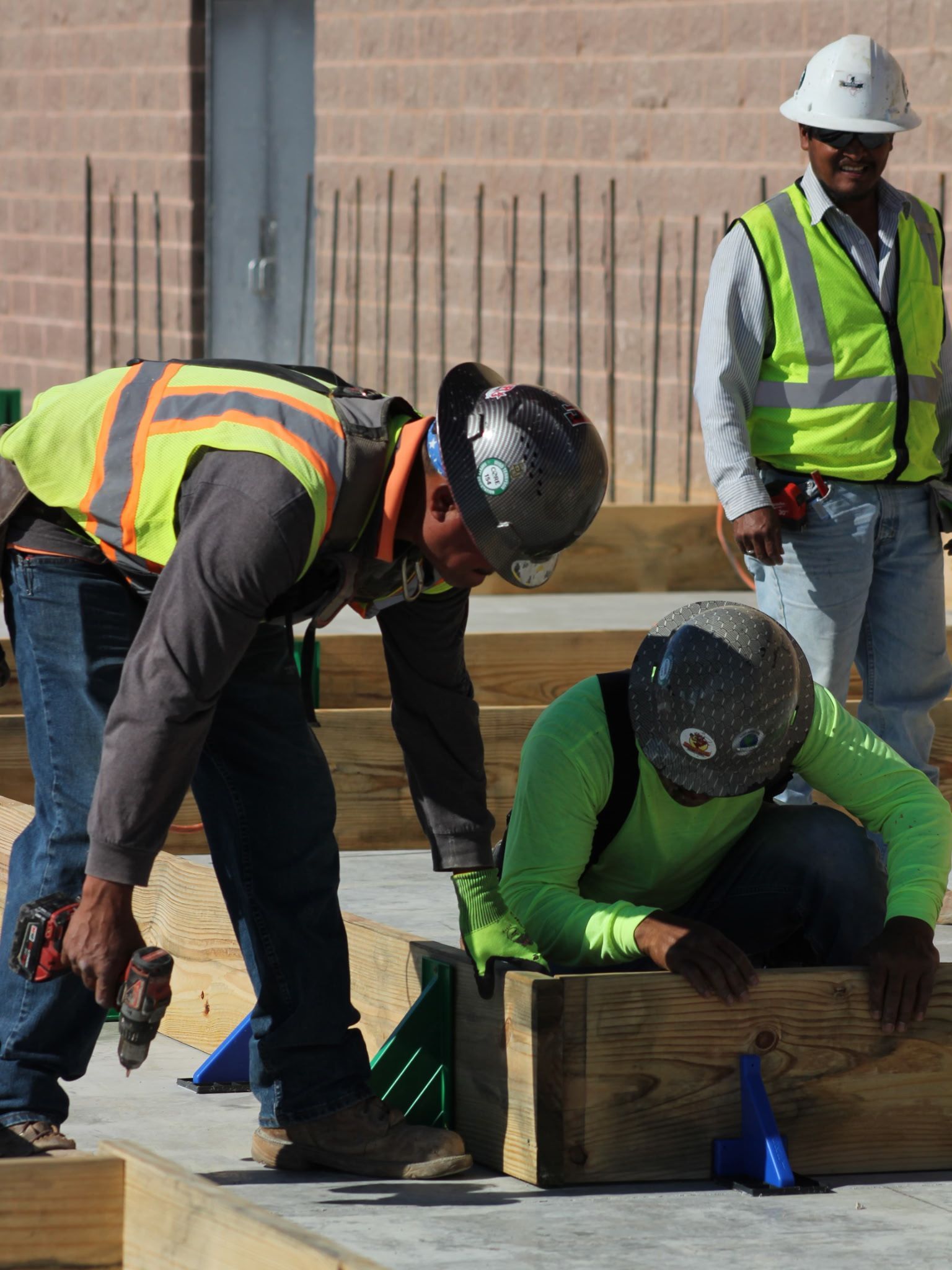 Construction workers in safety vests and helmets bent over timber at a work site.