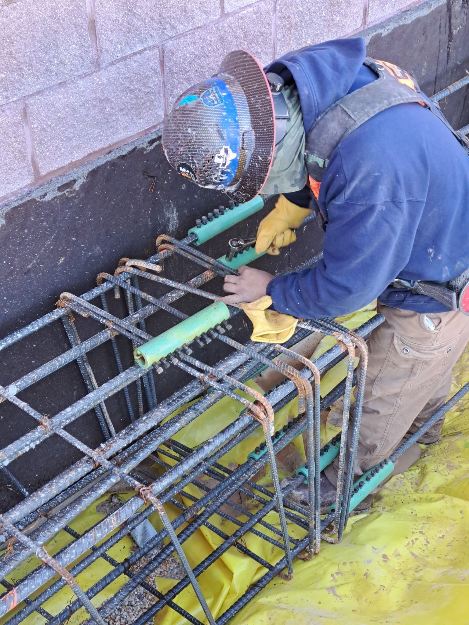 Worker welding rebar cage near a wall, wearing blue jacket, helmet, and yellow gloves.
