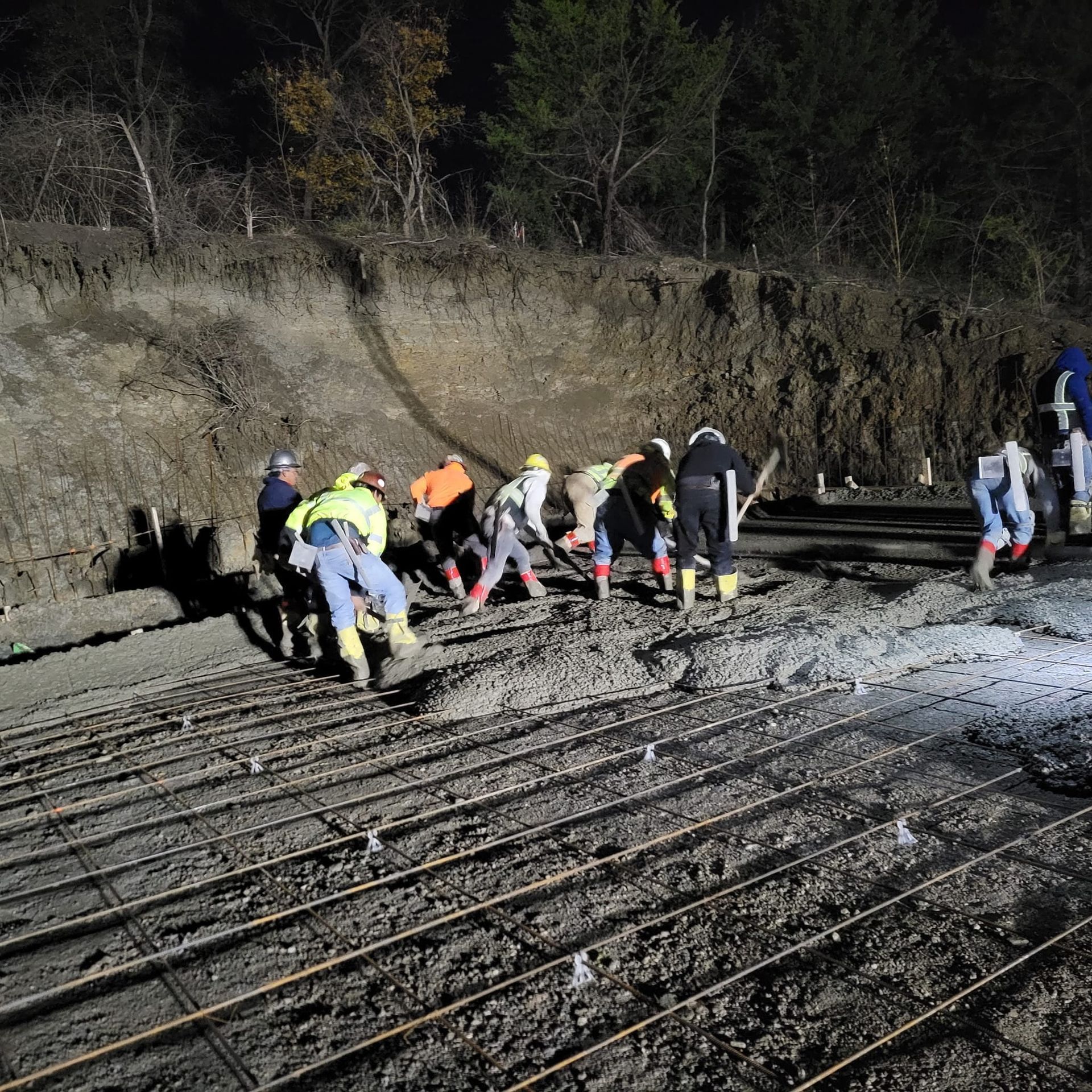 Workers in reflective gear laying rebar on a muddy construction site at night