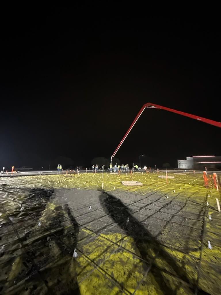 Nighttime construction site with a red concrete pump arm over a muddy, fenced ground area.
