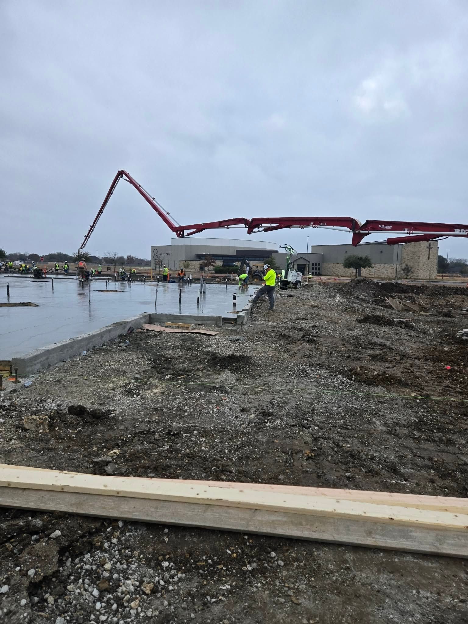 Construction site with a red concrete pump truck beside a muddy waterway under a cloudy sky
