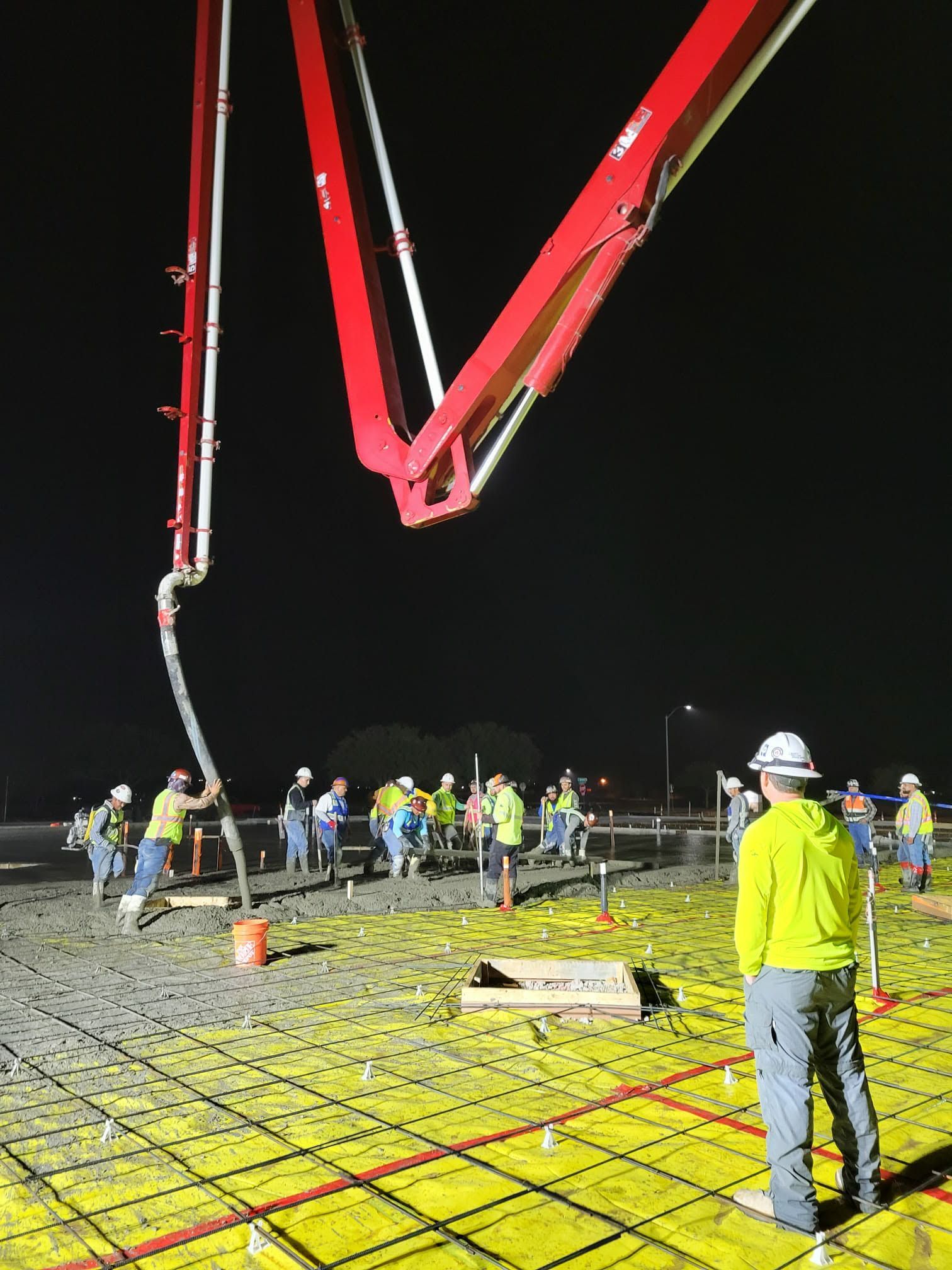 Workers gather beneath a red crane arm at a nighttime concrete pour on a construction site.