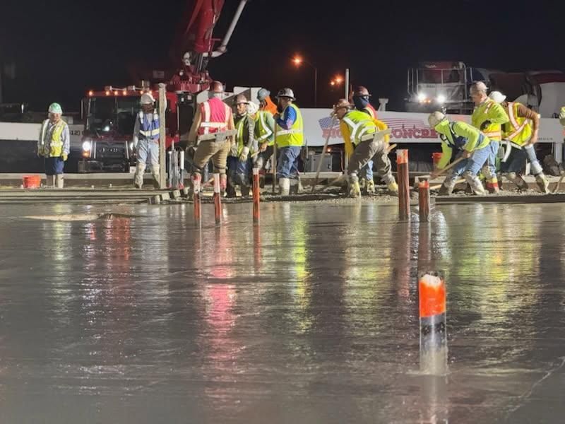 Construction workers pouring concrete at night on a wet road, with a cement truck and crane nearby