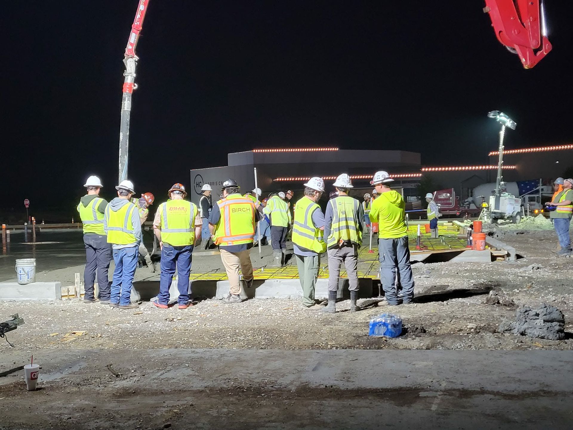 Construction workers in reflective vests stand at a night worksite beside concrete pumps and gravel.