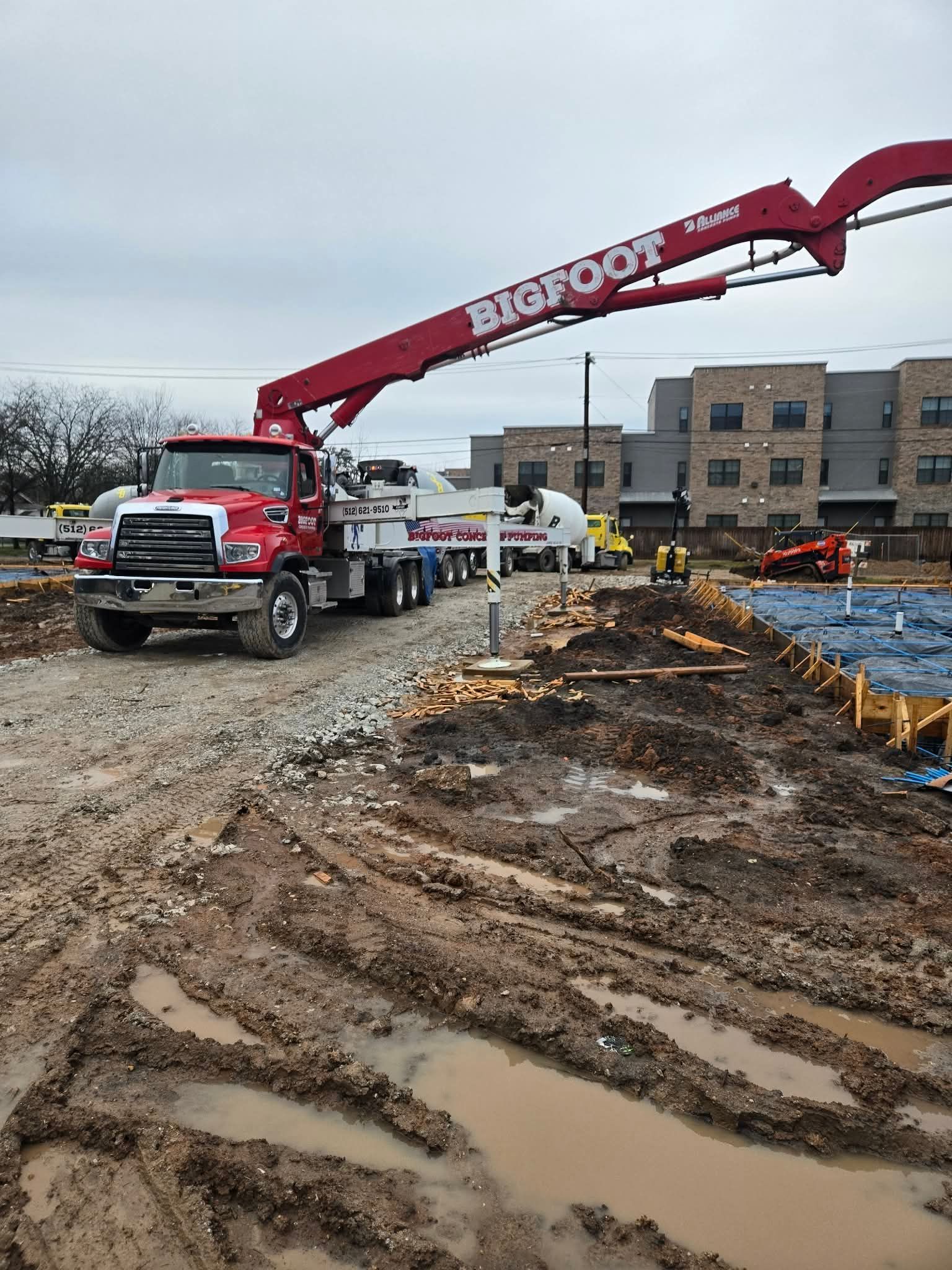 Red concrete pump truck pouring at a muddy construction site beside an unfinished brick building.