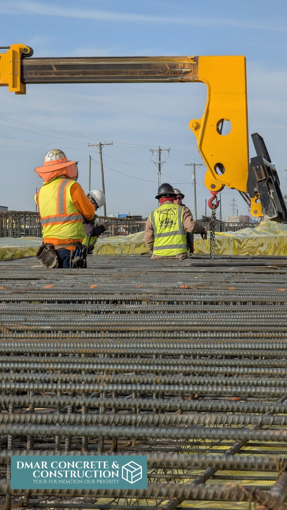 Construction workers seated beside heavy excavator arm at a roadwork site with stacked rebar in the foreground