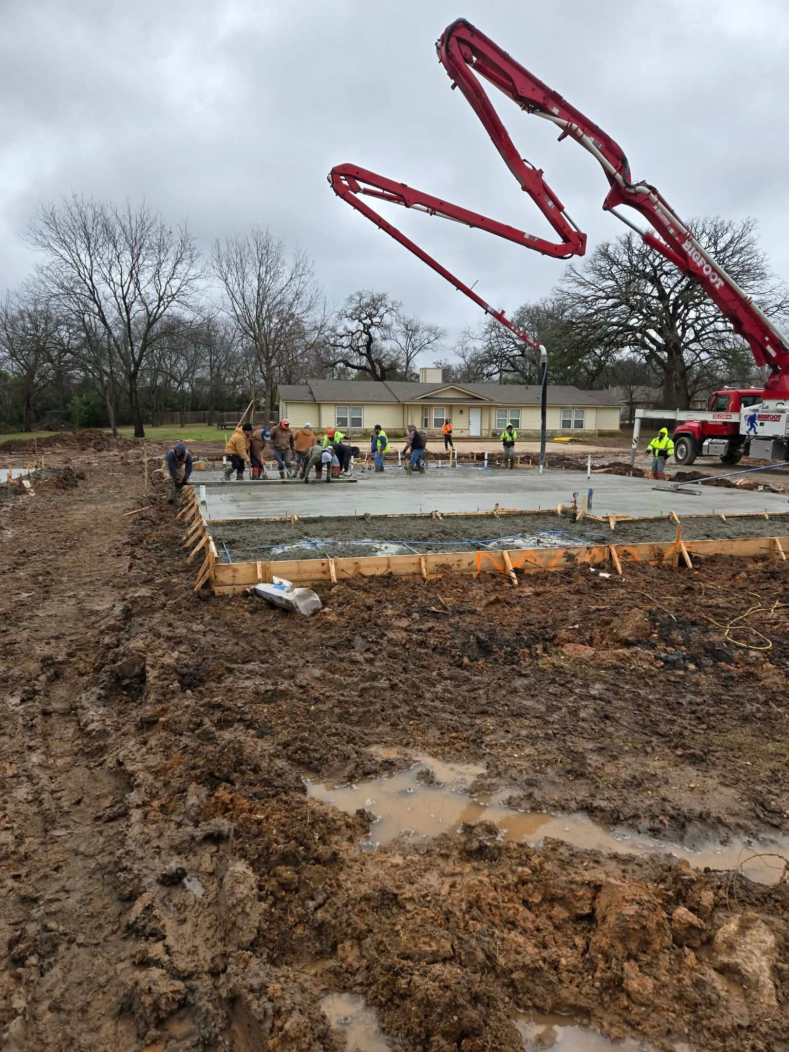 Concrete pool construction site with muddy ground, workers, and a red boom pump in the background