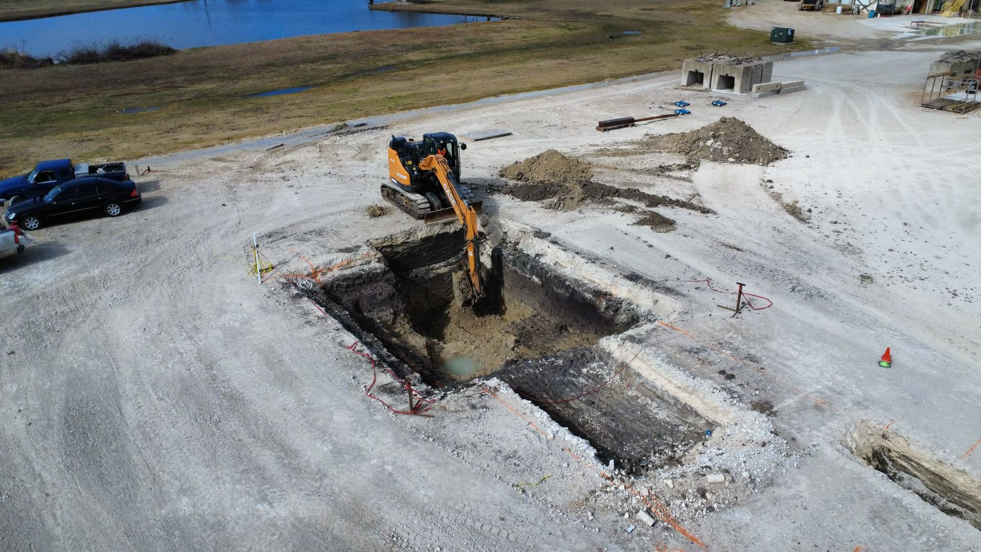 Excavator digging a deep trench in a sandy construction site near water, with vehicles nearby