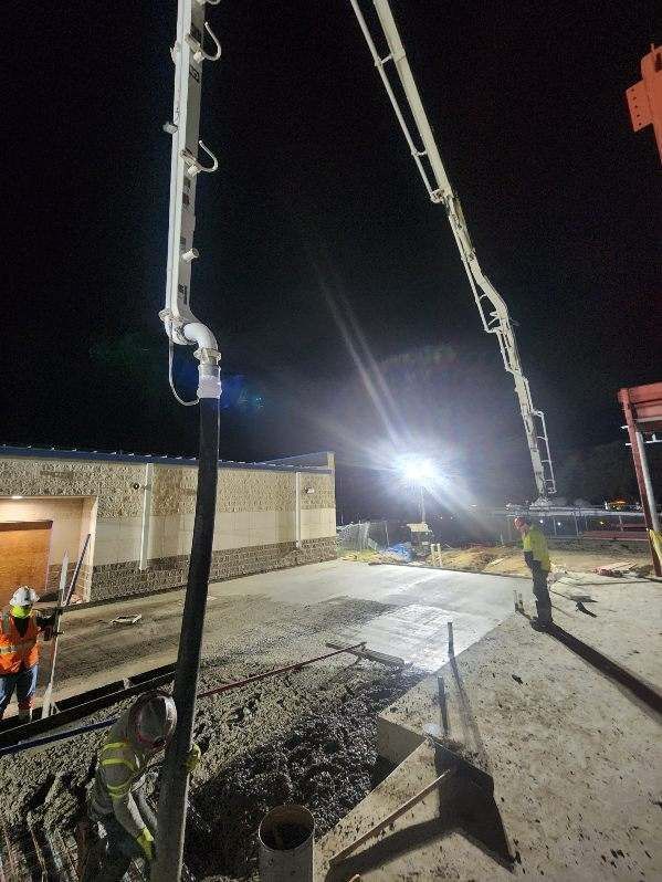 Night construction site with two cranes, a bright floodlight, and workers near a concrete slab.