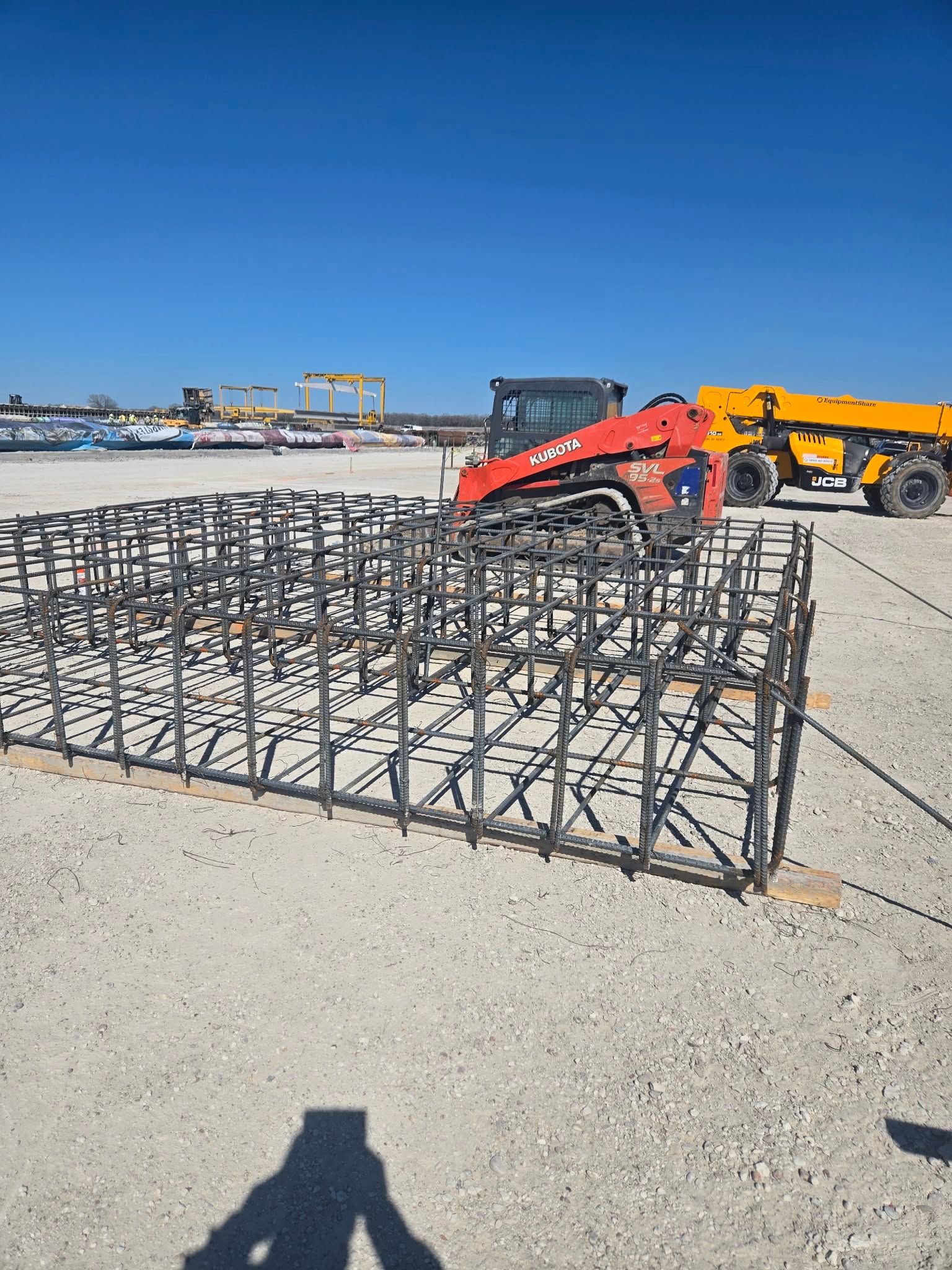 Construction site with red and yellow machinery moving metal rebar cages on a sandy surface