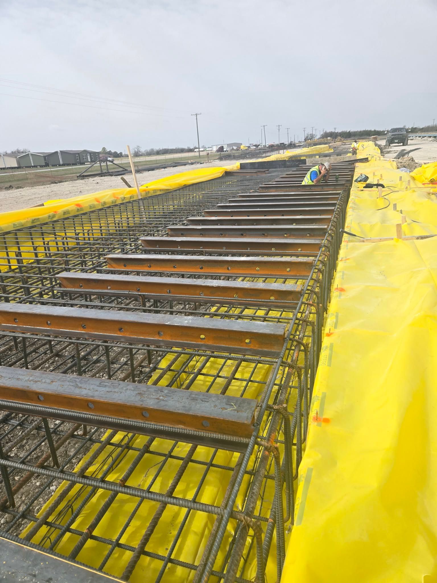 Steel rebar framework on a construction site, covered with yellow tarps along a road or embankment.