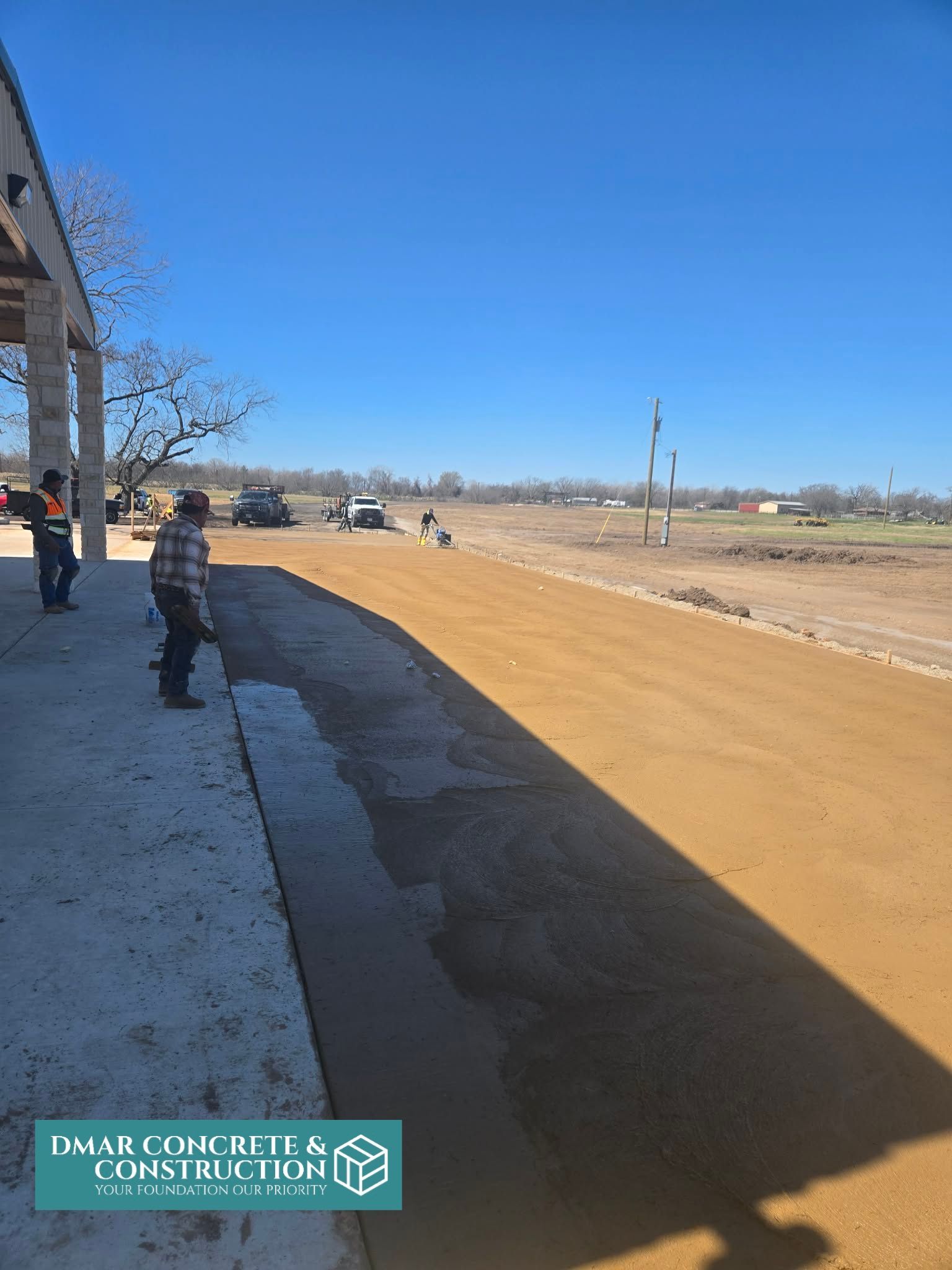 Sidewalk beside a dirt road under clear blue sky, with a few people walking near a building.