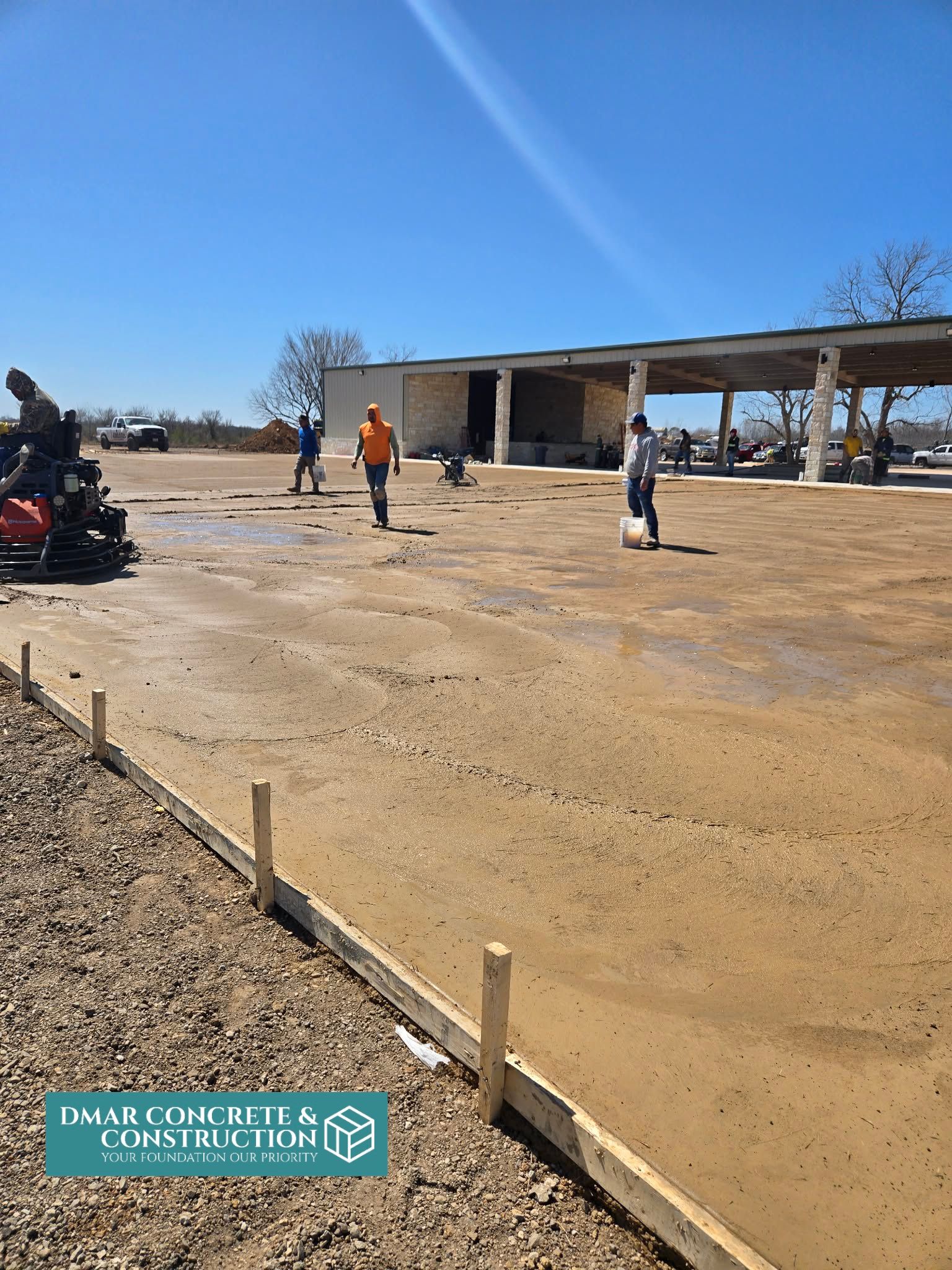 Workers on a dusty construction site beside a partially built metal-roof structure under a clear blue sky