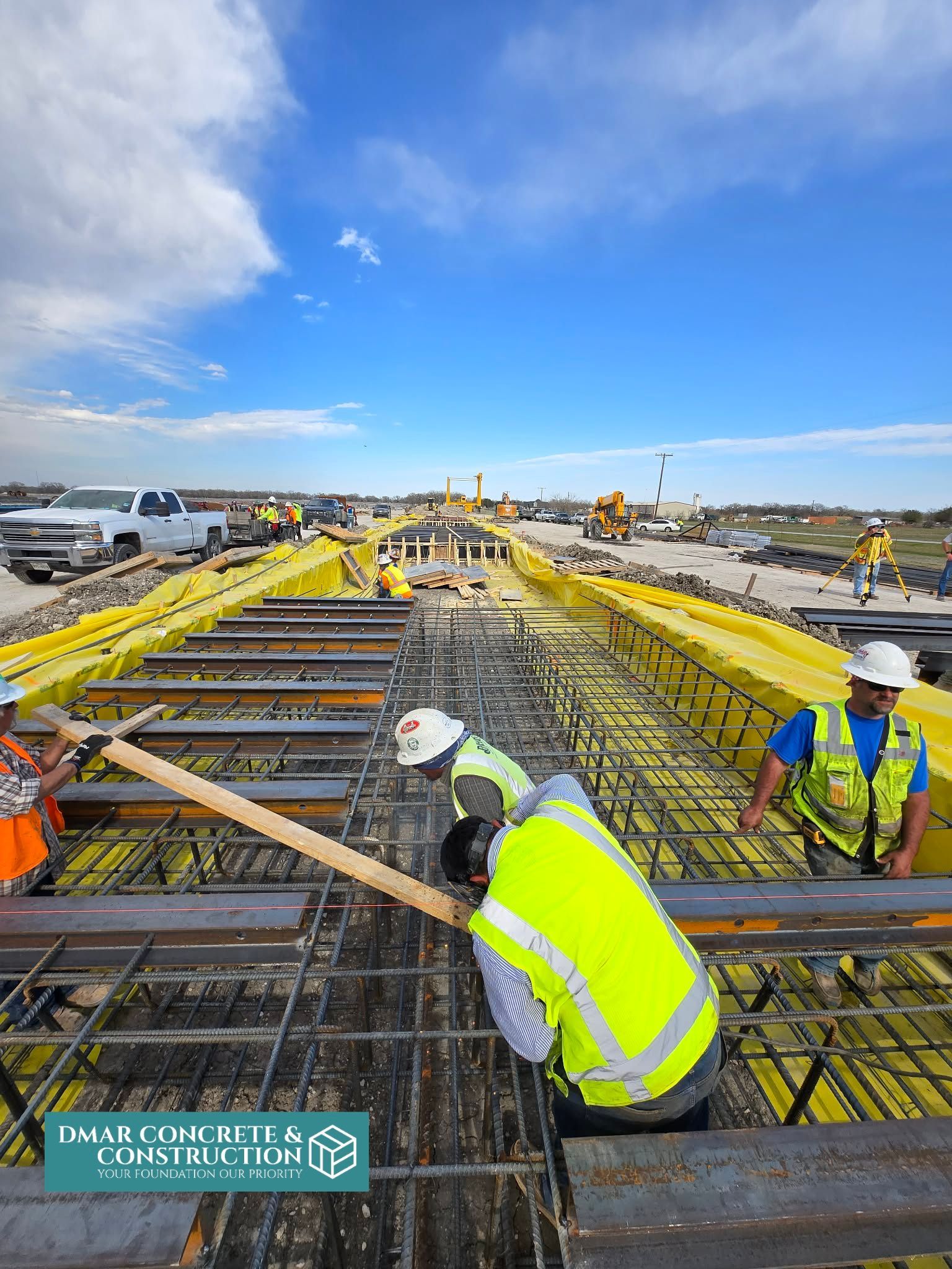 Construction workers in high-visibility vests pour concrete on a bridge deck under a blue sky