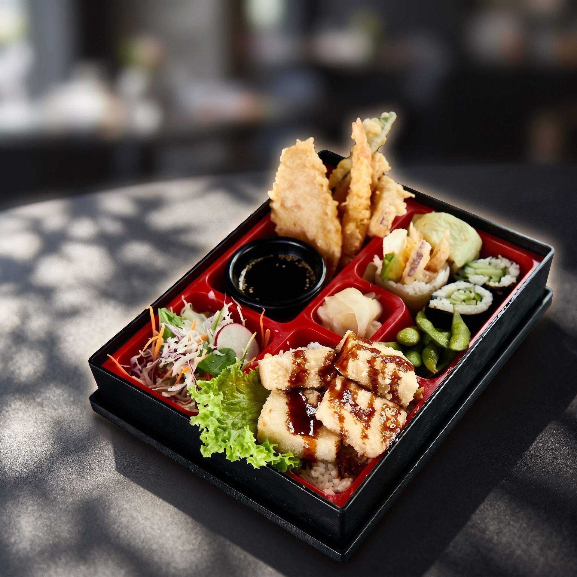 Black bento box with assorted sushi, tempura, salad, and dipping sauce on a table