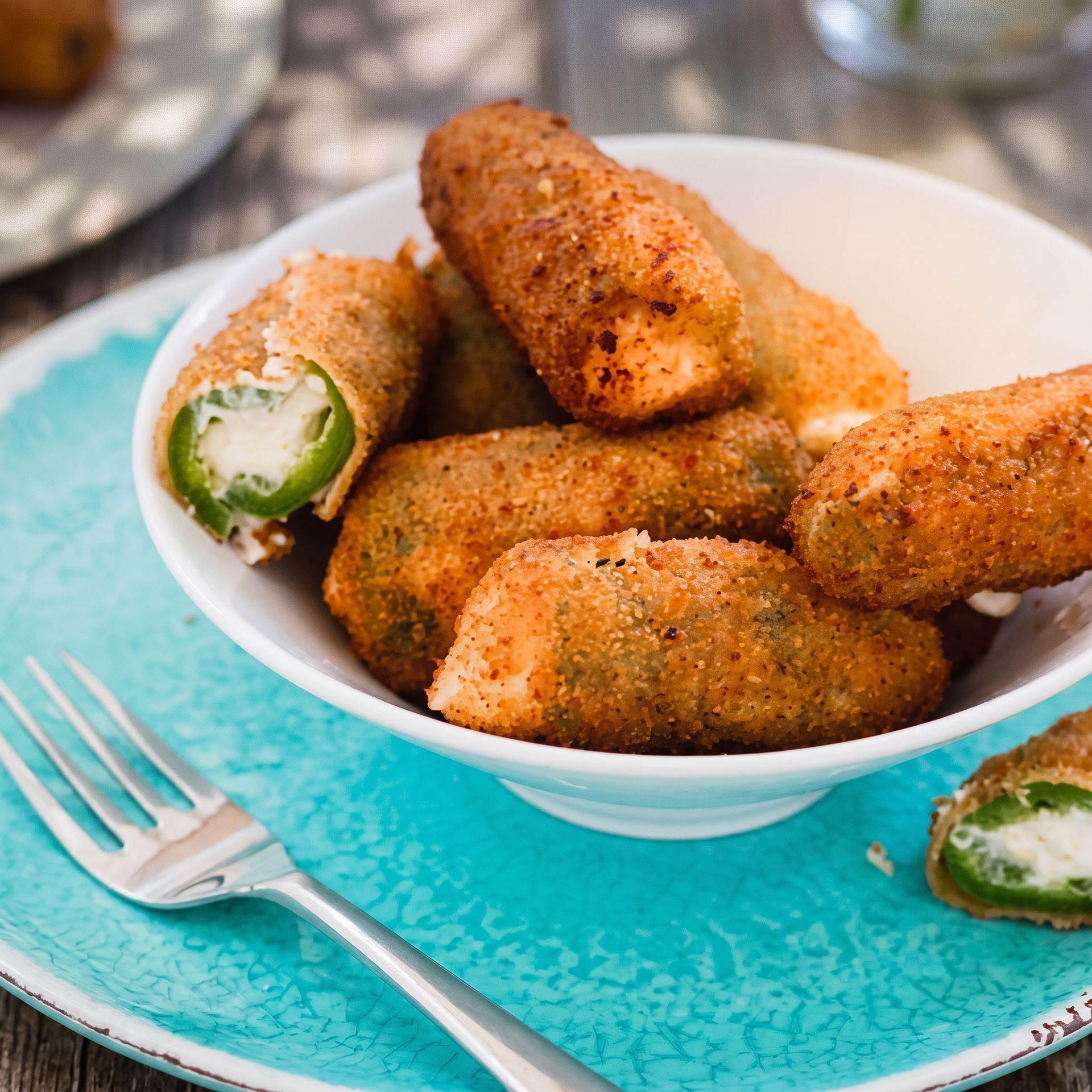Breaded jalapeño poppers in a white bowl on a turquoise plate with a fork