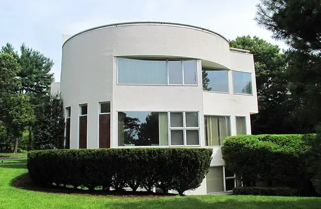 A white house with a lot of windows is surrounded by bushes and trees