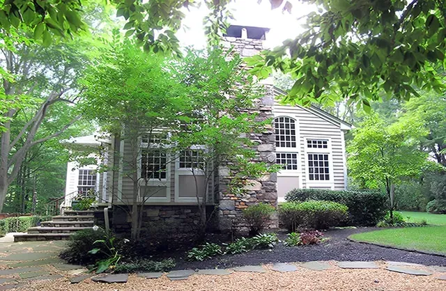A house with a stone chimney is surrounded by trees and gravel