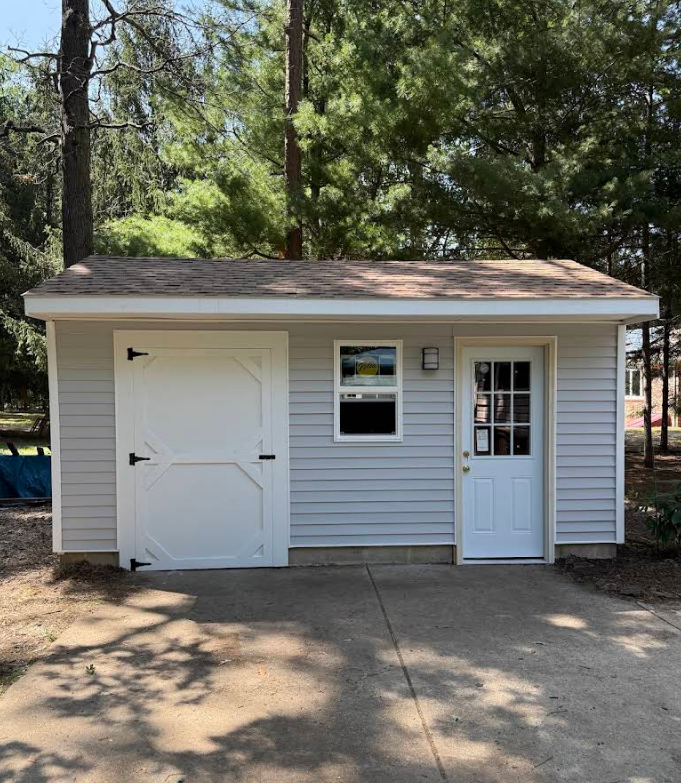 Small white shed with double doors and a side entry door in a wooded yard