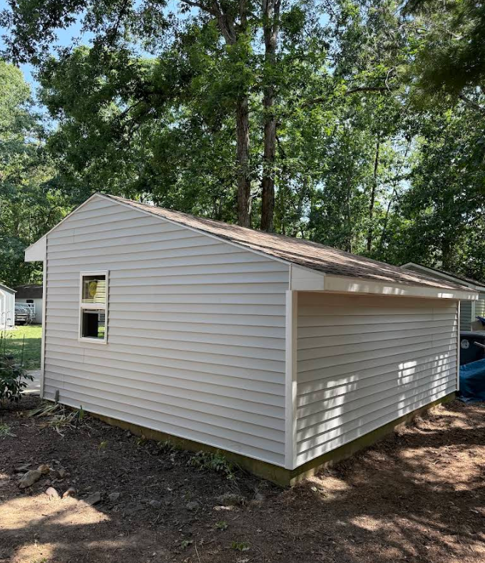 White vinyl-sided detached garage with a tan shingle roof, set among trees in a wooded yard.