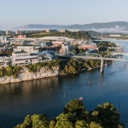 Riverfront cityscape with a bridge spanning a wide blue river, hills in the background, and buildings along the shore