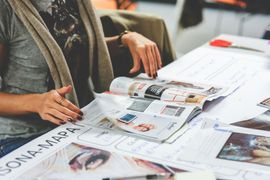 Two people reviewing newspapers and magazines spread across a table in a bright room