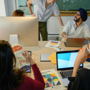 People collaborating around a conference table with laptops, papers, and a large monitor in a meeting room