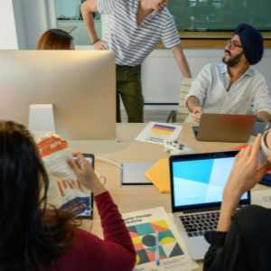 People collaborating around a conference table with laptops, papers, and a large monitor in a meeting room