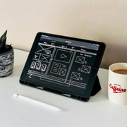 Desk with a tablet displaying a design layout, a small potted plant, and a coffee mug on a white surface.