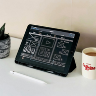 Desk with a tablet displaying a design layout, a small potted plant, and a coffee mug on a white surface.