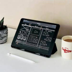 Desk with a tablet displaying a design layout, a small potted plant, and a coffee mug on a white surface.