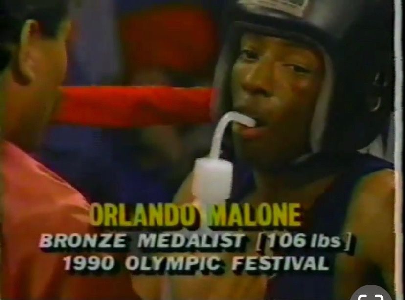 Boxer Orlando Malone in headgear, resting with a mouthguard, captioned “Bronze Medalist 106 lbs, 1990 Olympic Festival”