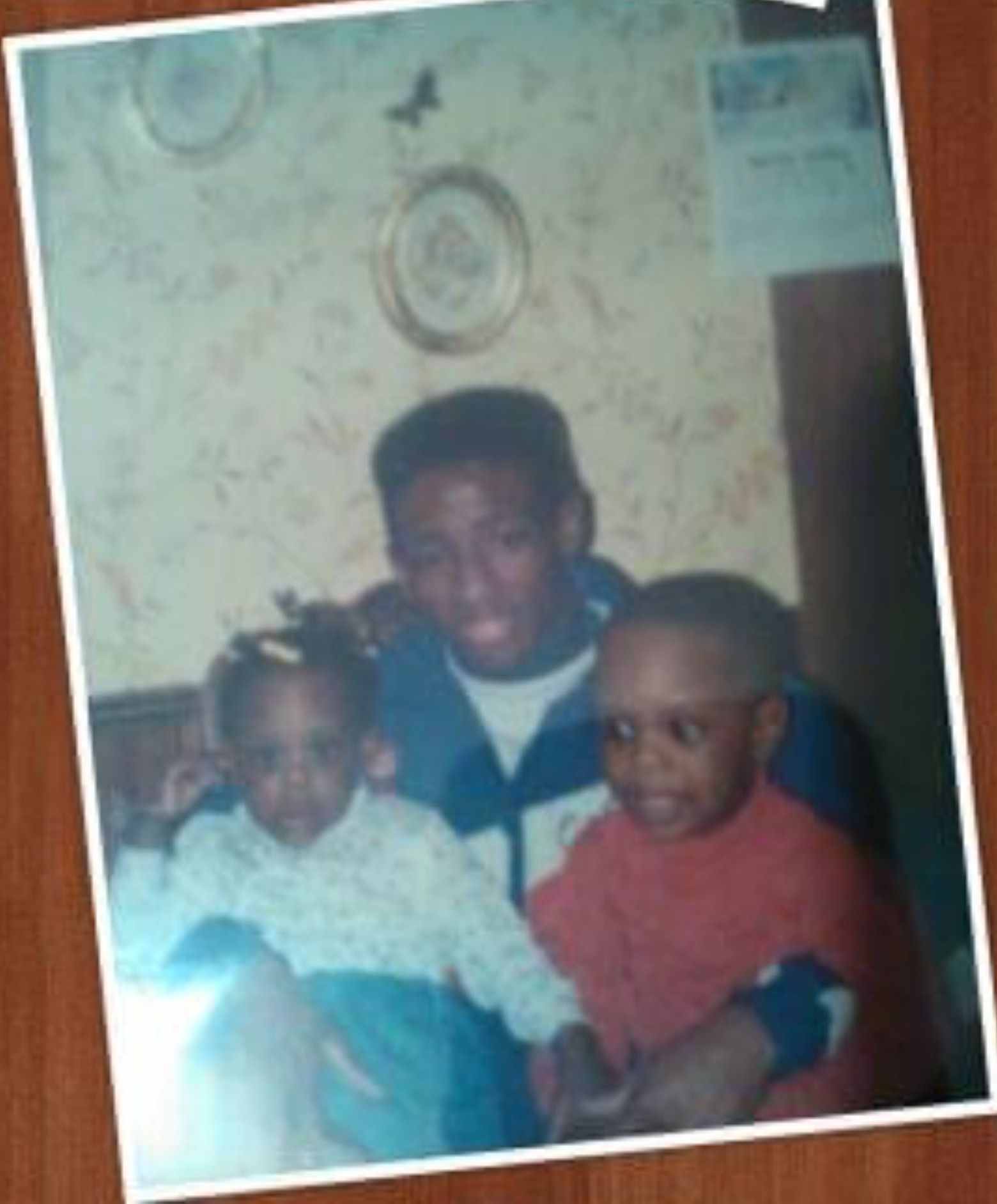 Three children sitting together indoors, with a wallpapered wall and framed items behind them.