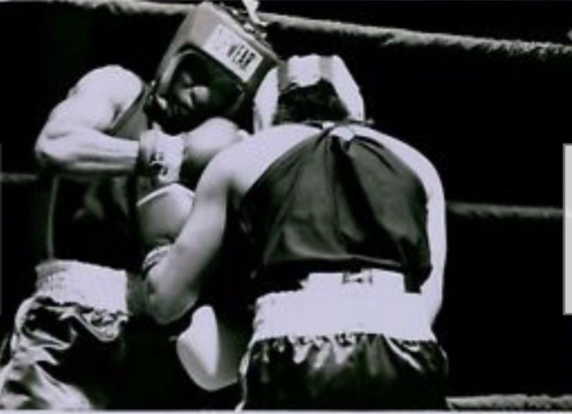 Two boxers in gloves and headgear clinch during a match in a boxing ring.