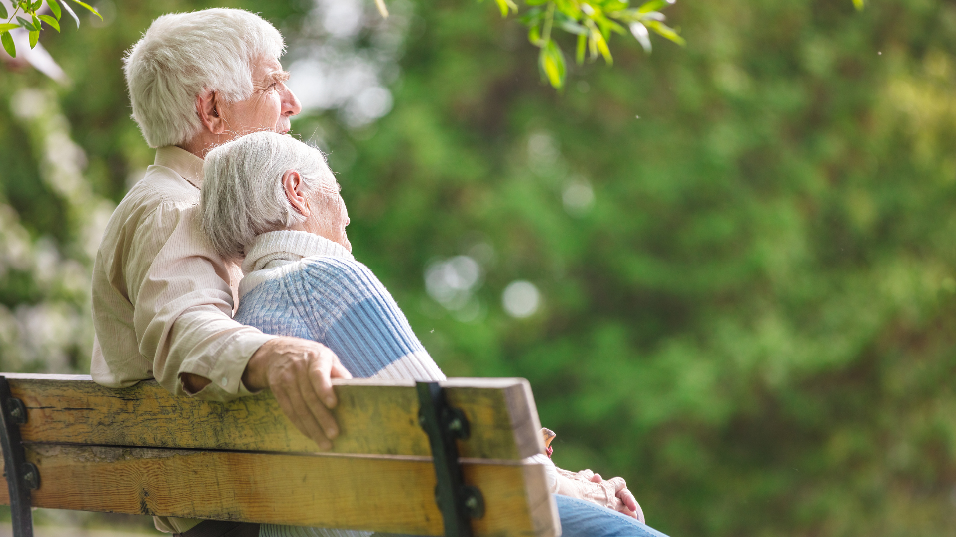 Two elderly people sitting on a wooden bench in a green park, facing away from the camera.