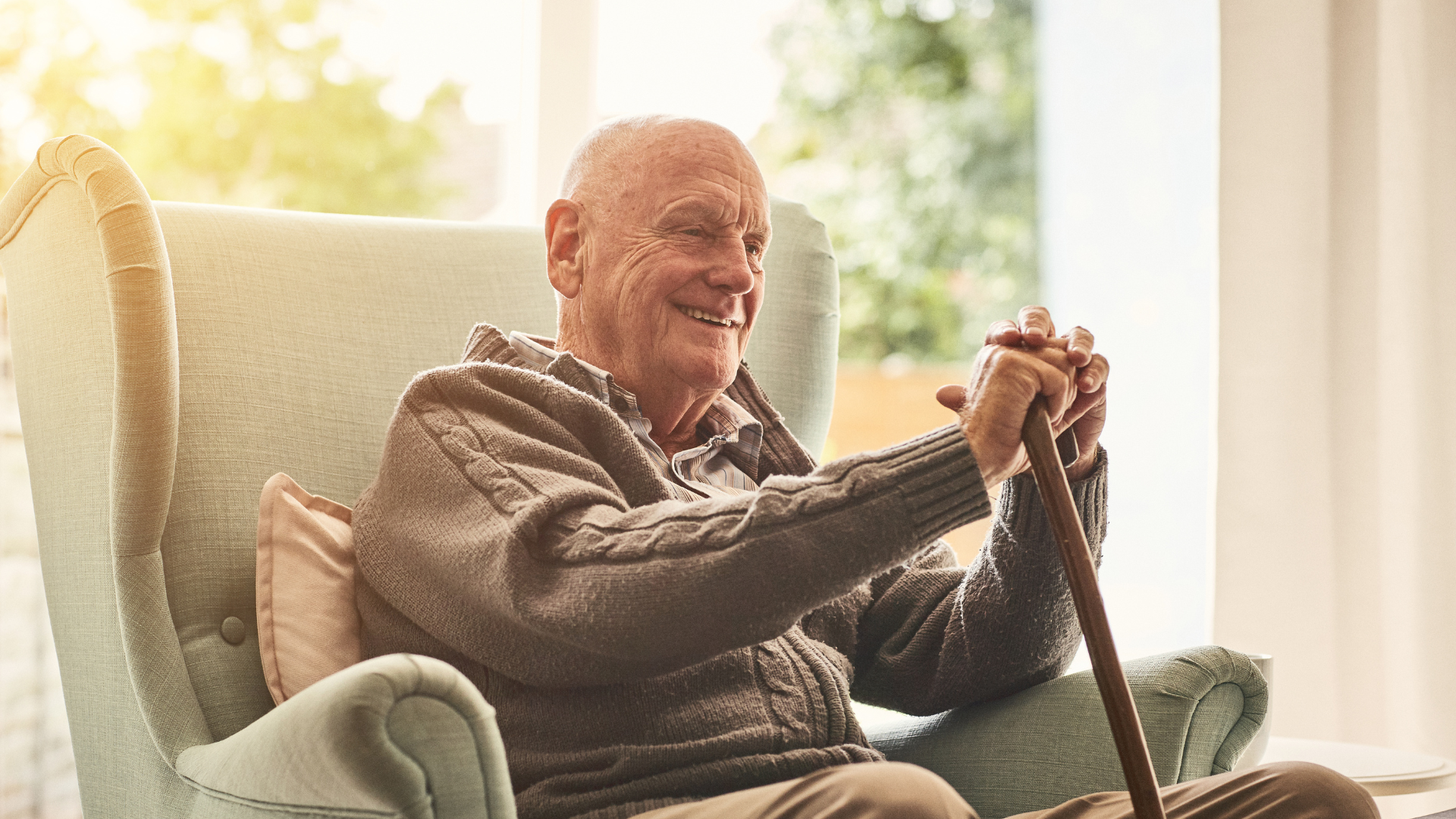 Smiling man sits in an armchair by a sunlit window, holding a cane and relaxing at home