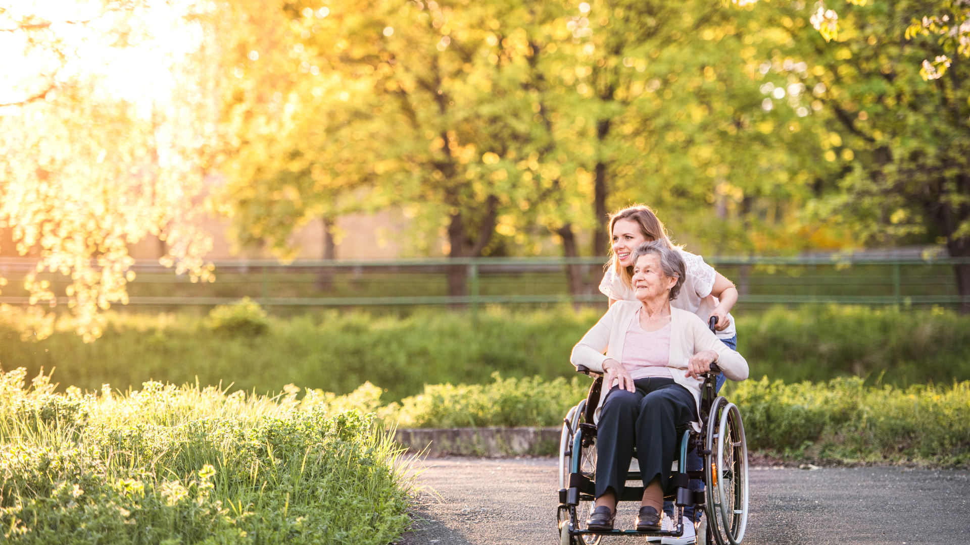 Two women in a wheelchair on a sunlit park path, with one pushing the other among green hedges.
