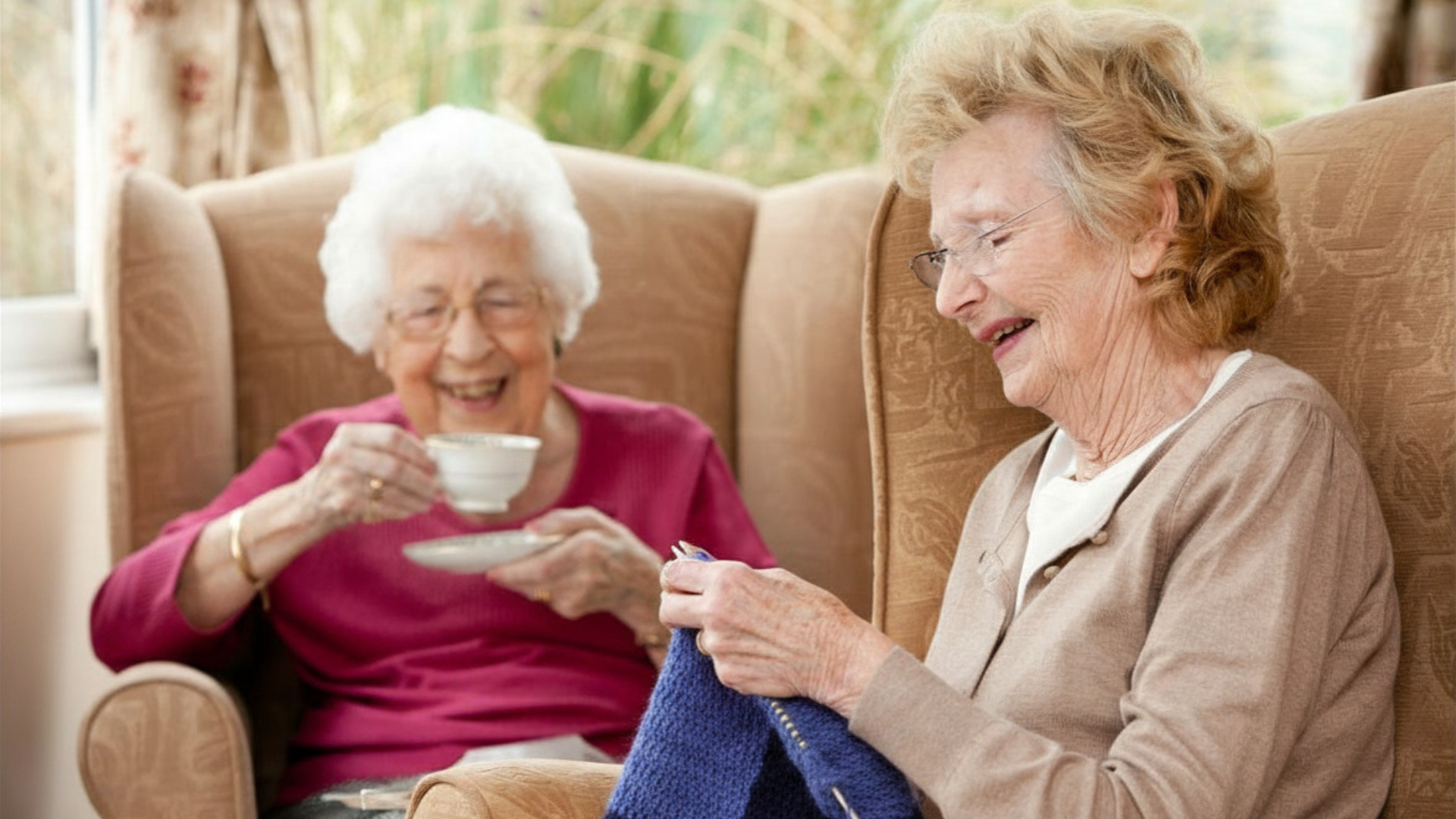 Two people relaxing on a couch, smiling and drinking tea while knitting in a bright living room.