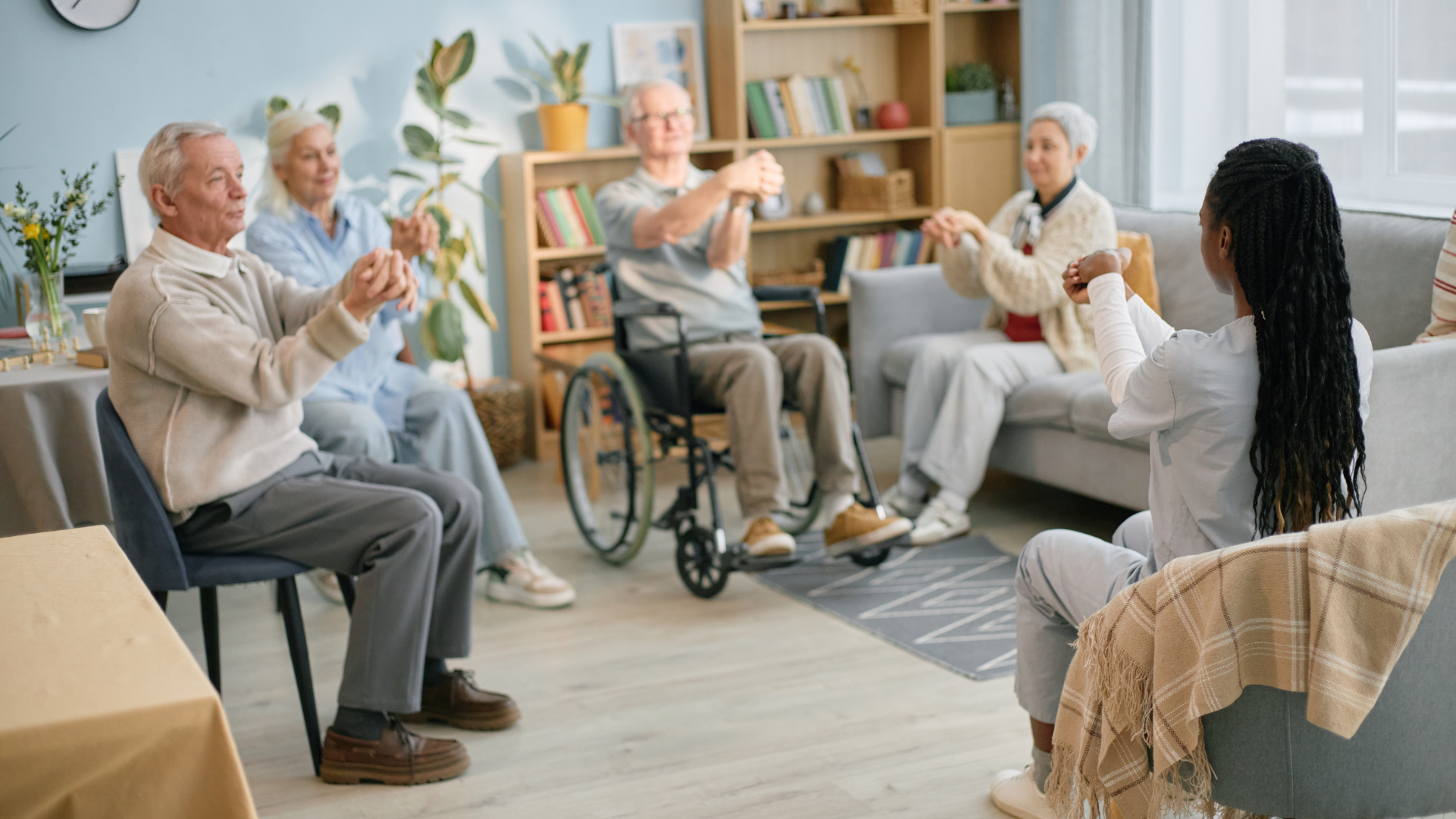 Group of seniors doing seated exercises in a bright living room with a coach leading them.