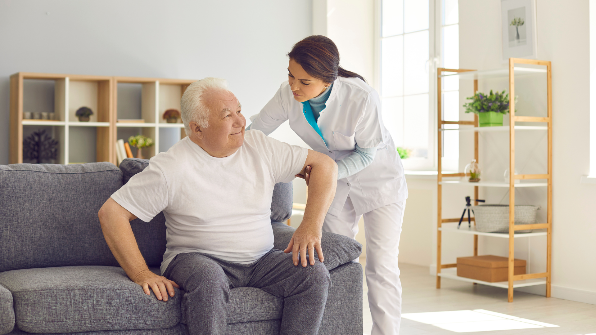 Caregiver assisting a seated patient in a bright living room beside a gray sofa